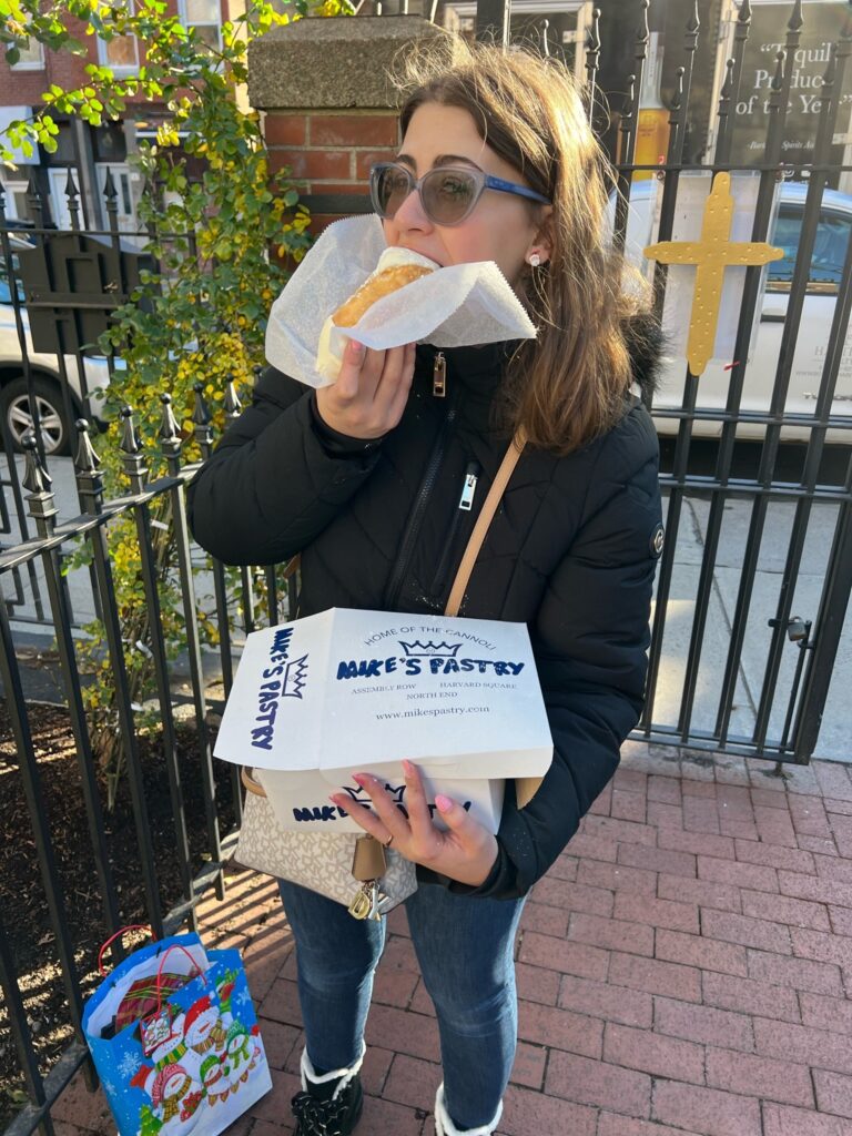 A woman eats a cannoli while holding a box of pastries from Mike's Pastry.