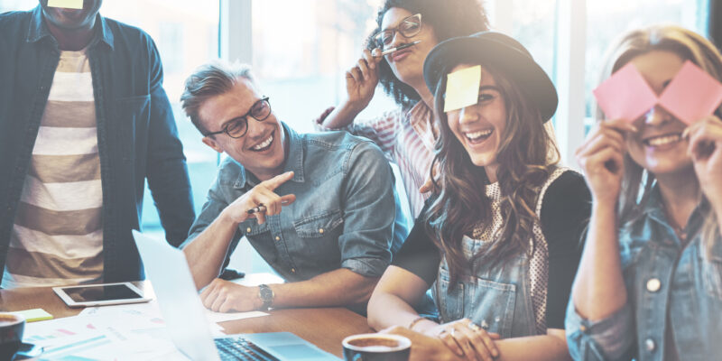 Joking group of diverse young adult coworkers playing with sticky notes on their faces as a distraction during a meeting