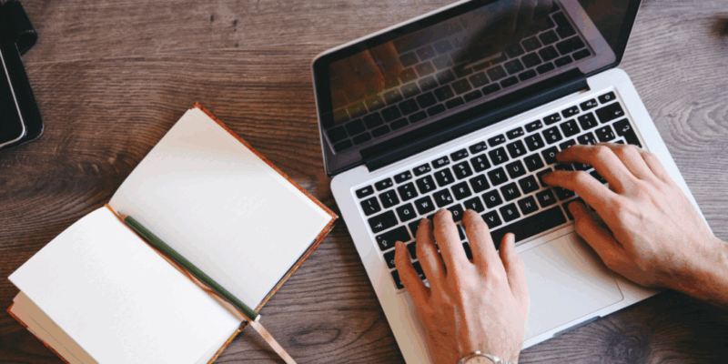 An overhead view of a man typing on his laptop, a gold watch on his left wrist. A notebook and pen open to his left with blank pages.