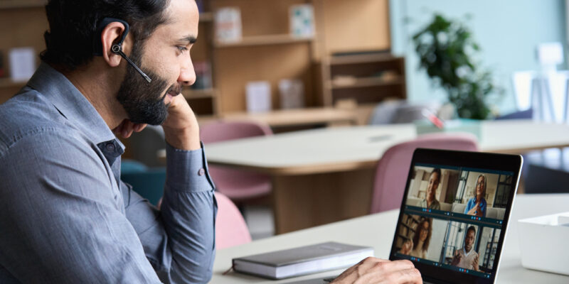 Indian business man wearing headset having virtual team meeting on video conference call using laptop work from home office talking to diverse people group in remote teamwork online distance chat.
