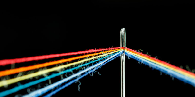 multi-colored threads for sewing in the form of a rainbow pass through an antique needle on a black background close-up